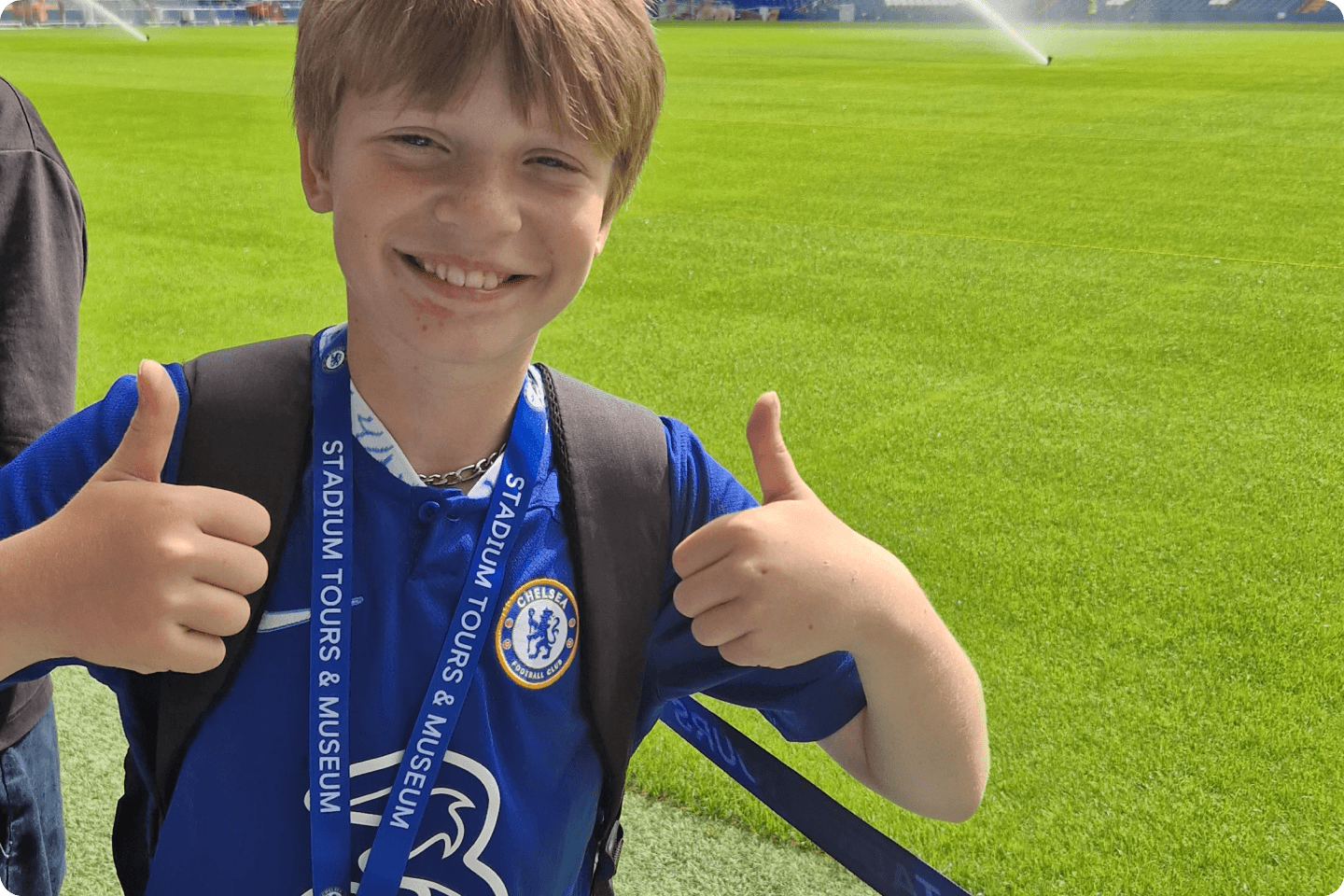 A smiling child gives two thumbs up and wears a blue sports jersey with a circular badge on the chest. The child is also wearing a blue lanyard around their neck with the text "STADIUM TOURS & MUSEUM." They stand on a lush green field with sprinklers in the background.