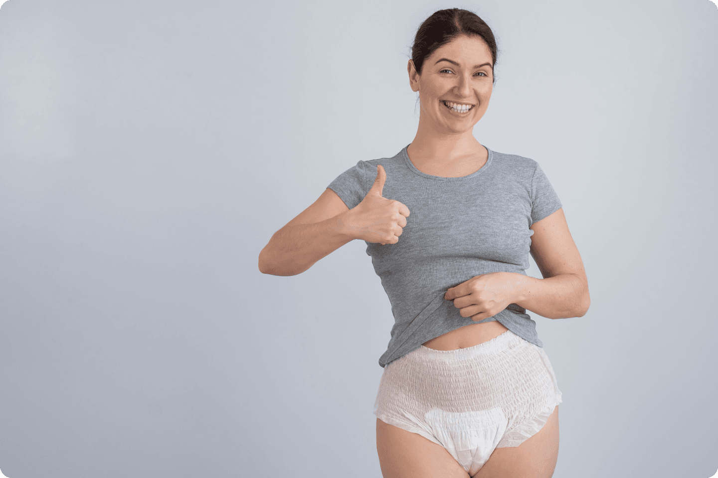 A smiling woman in a grey t-shirt and adult nappy gives a thumbs-up gesture. She holds up a corner of her shirt slightly to reveal the waistband of the nappy. The background is plain and light-coloured.