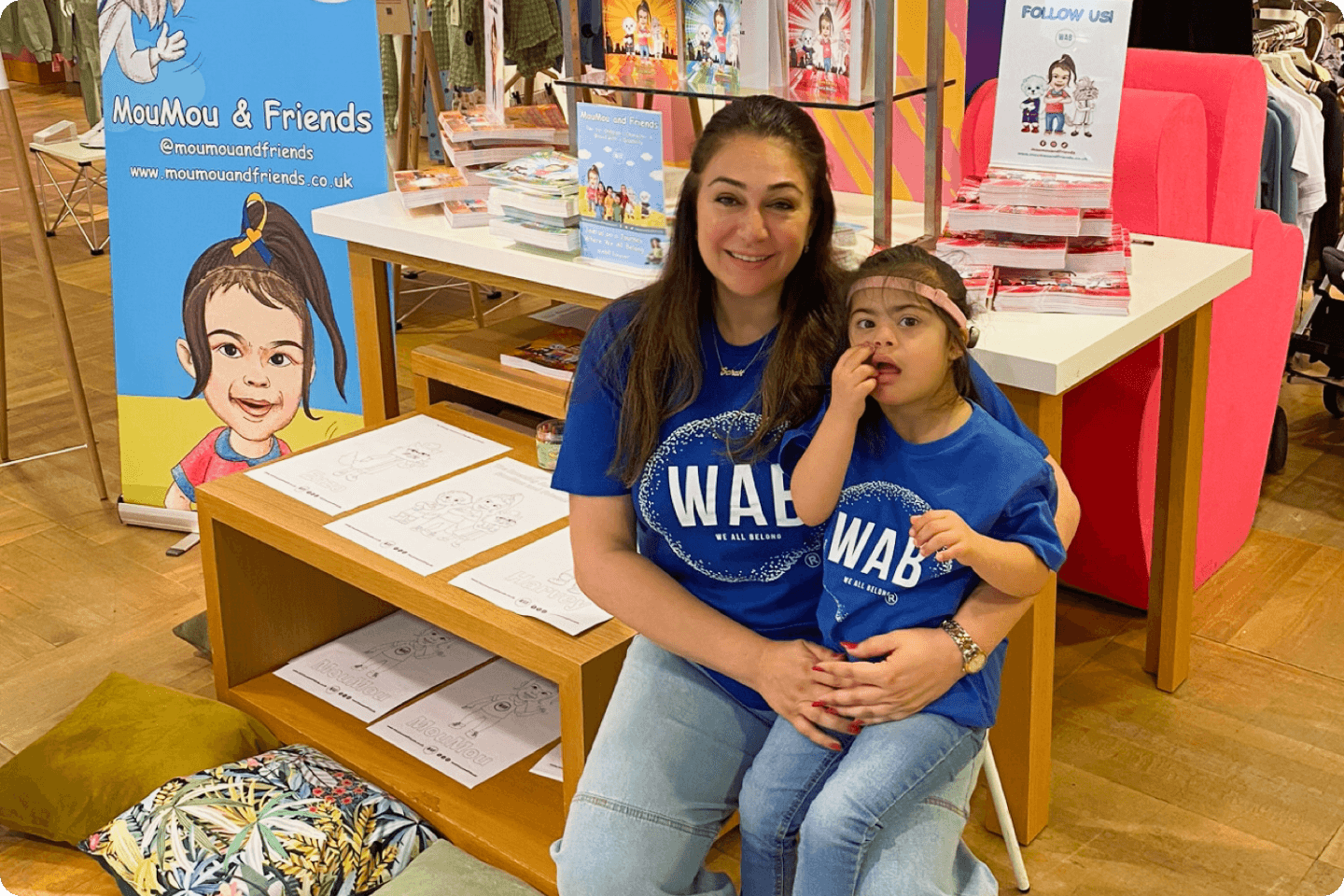 Amani is sitting on Sara's lap, and they are both smiling. The MouMou and Friends brand is visible behind them. They are both wearing royal blue WAB® t-shirts.