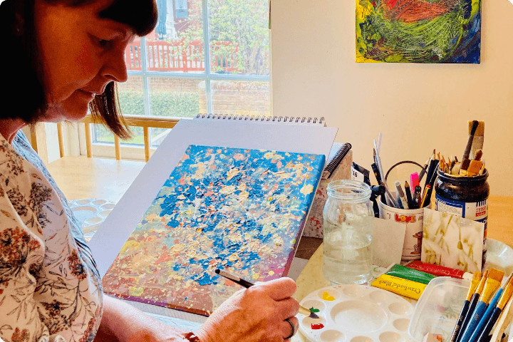 A woman paints a colourful abstract piece on a table with various art supplies. Sunlight streams through a window, illuminating her workspace.