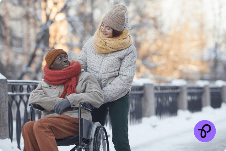 A couple in the snow, wrapped up warm, a black man is a wheelchair user and wears an orange scarf, a white woman wears a yellow scarf and is pushing the wheelchair for a post about keeping warm for disabled people