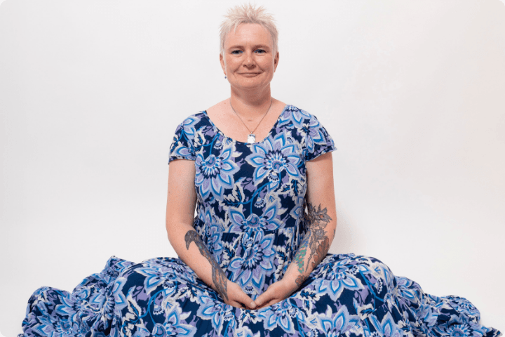 A woman with short blonde spiky hair sits with a content gentle smile on her face, she is wearing a blue floral long dress that covers her legs and flows onto the floor and she has her hands in her lap, there are tattoos on her arms