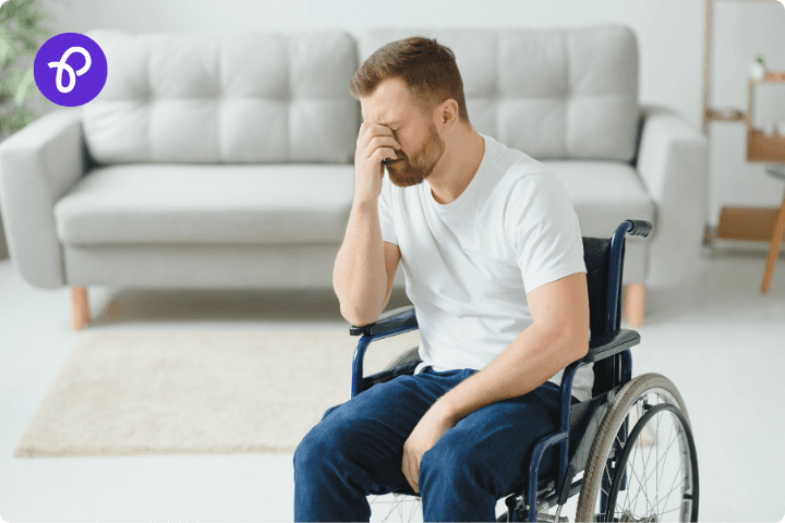 A white man is sat in a wheelchair, he wears white t-shirt and blue jeans and has a short beard, he has his head in his hand and looks stressed, behind him is a living room with a white sofa