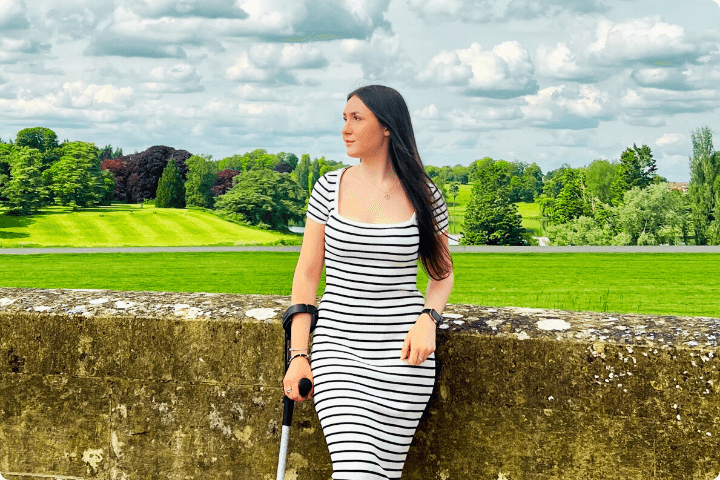 A young woman with long brunette hair is leaning against a stone wall with green landscape behind. She is wearing a black-and-white striped dress, holding a silver, glittery crutch on one side and is looking over into the distance.