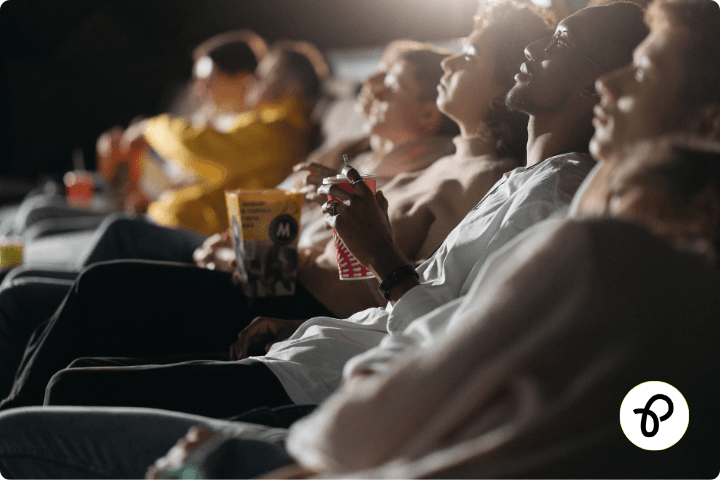 A moody dark image of a line of people sat in a movie theatre, their faces are lit by the light of a cinema screen