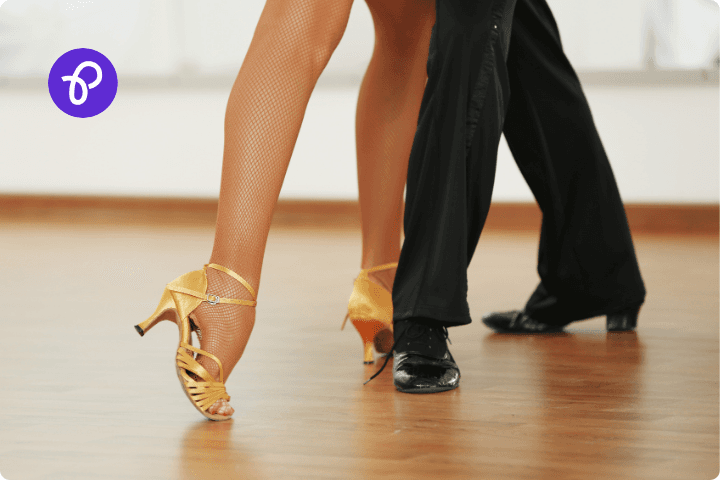 A male and female set of legs on a dance floor, the women's legs are bare and wearing gold shoes and the mens legs are wearing formal black trousers and dance shoes