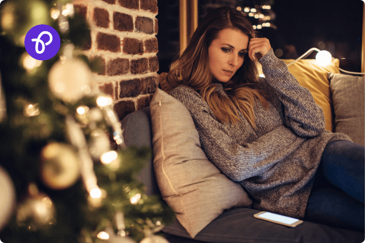 A white woman is sat on a sofa with a Christmas tree in the foreground, she has her hand to her head and looks anxious and sad, she is looking at a mobile phone