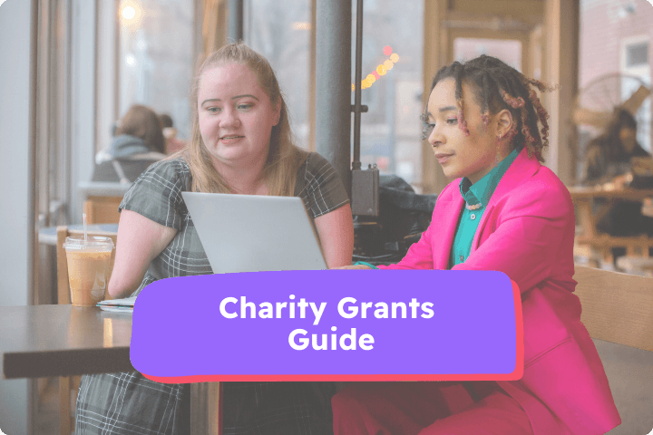 A white woman with a limb difference is sat at a table with a black woman, they are both looking at a laptop. the text reads Charity Grants Guide