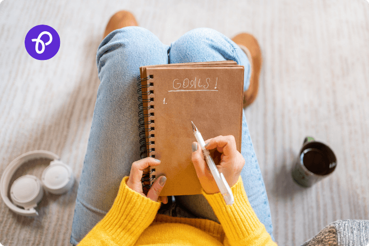 An overhead view of a woman wearing jeans and a yellow jumper sat with a notebook and pen in her lap, she has written the word goals