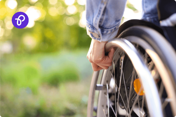 A close up of the left wheel of a wheelchair with a hand resting on the rim, the person is wearing a blue denim jacket, there is the Purpl disabled discounts logo in the corner.