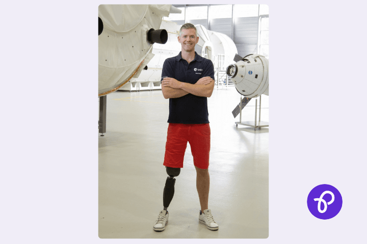 A white man, John McFall who is the first disabled man to go into space, he has short brown hair, wears a blue polo shirt and red shorts, he has a lower limb difference and has a prothetic leg and is smiling whilst stood in an aircraft hangar with airplanes behind him