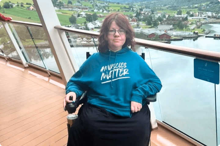 Sara Stacey is a white woman with shoulder length brown hair, she is wearing a teal jumper that says Muscles Matter on the front and is a wheelchair user, she is on a balcony with a view of water and a mountain town in the background