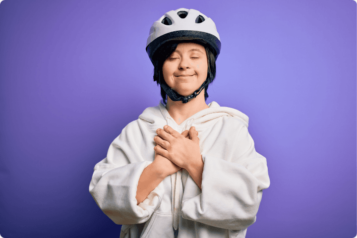 a purple background with a white person with Down's syndrome in a white hoodie and bicycle helmet smiling with their eyes closed and hands clasped on their chest