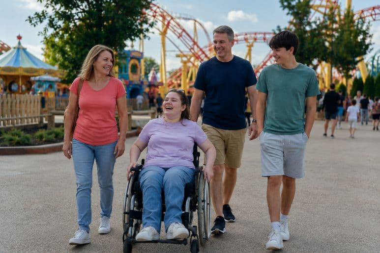 Family enjoying free carer entry at a theme park with a young disabled adult using a wheelchair.