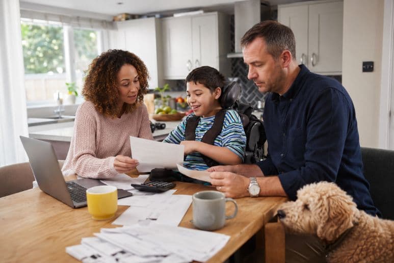 Family with a disabled child in a wheelchair reviewing household bills at a kitchen table with their pet dog nearby, showing the extra cost of disability for UK households.