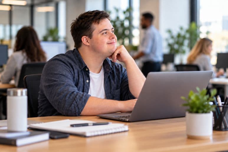 Man with Down syndrome working on a laptop in a modern office with colleagues in the background.